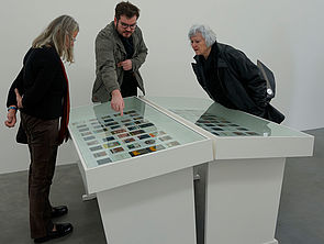 Niklas Washausen zeigt auf ein Foto in einer von zwei Vitrinen. Zwei Frauen beugen sich neugierig darüber.