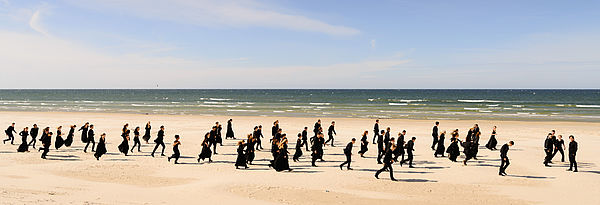 Ungefähr 60 Frauen und Männer vom Baltic Sea Philharmonic laufen am Ostseestrand entlang. Sie sind alle in Schwarz gekleidet. Hinter ihnen liegt die Ostsee.
