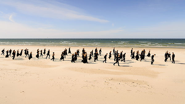 Ungefähr 60 Frauen und Männer vom Baltic Sea Philharmonic laufen am Ostseestrand entlang. Sie sind alle in Schwarz gekleidet. Hinter ihnen liegt die Ostsee.