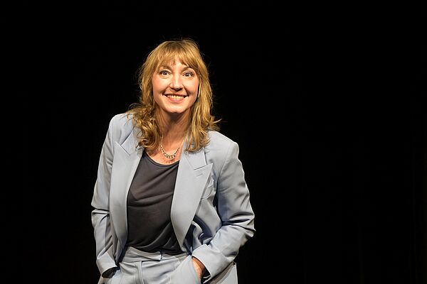 Johanna Falckner steht vor einem schwarzen Hintergrund. Sie trägt einen graue Hose und einen grauen Blazer, dazu ein dunkles Shirt. Ihre Hände stecken in den Hosentaschen. Sie lacht in die Kamera.