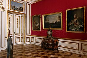 Blick in einen Ausstellungsraum. An den roten Wänden hängen Gemälde.