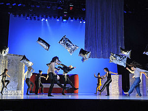 Auf einer Bühne schweben silberne Kissen. Neun Frauen und Männer tanzen. Auf der Bühne steht ein Sofa. 
