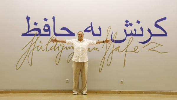 Günther Uecker steht mit ausgebreiteten Armen vor einem Schriftzug „Huldigung an Hafez”. Der Schriftzug steht in Deutsch und Persisch an der Wand.