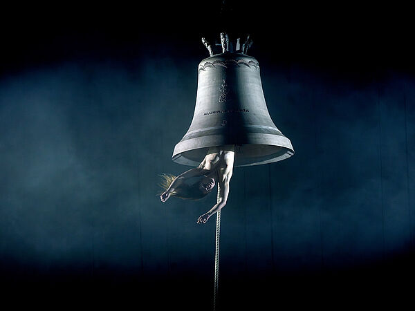 Eine große Kirchenglocke. Aus der Glocke hängt kopfüber eine Frau. Die Frau hängt bis zur Taille aus der Glocke heraus. Die Frau ist nackt.