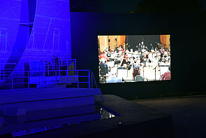 Auf einer Leinwand neben der Bühne spielt die Staatskapelle.