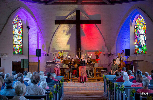 Eine kleine Kirche. Vor dem Altar stehen Musiker. Sie spielen Instrumente. Eine Frau singt. Vor der Band sitzen Zuschauer.  