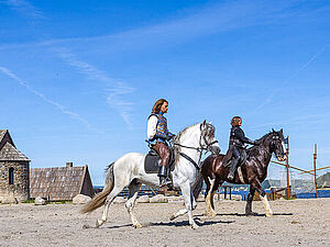 Zwei Darsteller reiten auf Pferden durch die Kulisse.