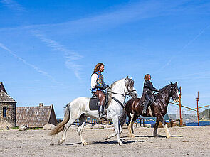 Zwei Darsteller reiten auf Pferden durch die Kulisse.