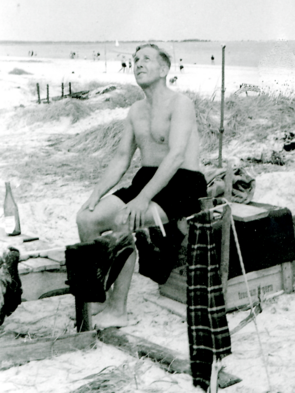 Ein Mann in Badehose sitzt auf einer Kiste am Strand.