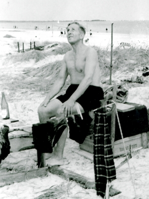 Ein Mann in Badehose sitzt auf einer Kiste am Strand.