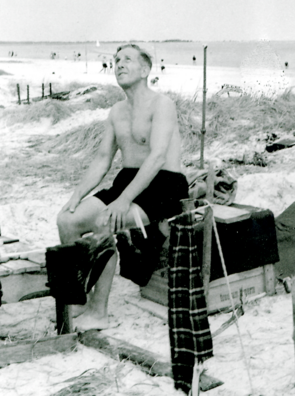 Ein Mann in Badehose sitzt auf einer Kiste am Strand.