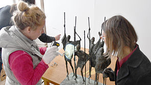 Zwei Frauen stehen vor Metallskulpturen. Eine von ihnen leuchtet mit einer Taschenlampe auf eine Figur.