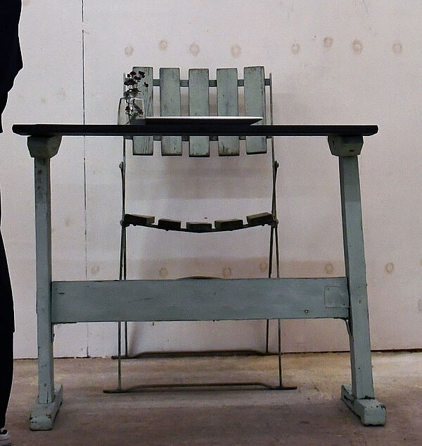 Ein Tisch und ein Stuhl vor einer weißen Wand. Darüber hängt ein Bild: Eine Person sitzt auf dem Stuhl am Tisch.