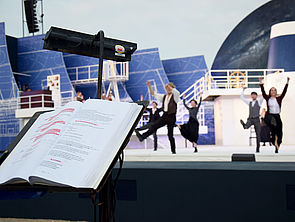 Auf einem Ständer liegt ein aufgeschlagenes Buch. Darin steht der Text zum Stück. Einige Passagen sind rot markiert. Im Hintergrund bewegen sich Schauspieler auf der Bühne.