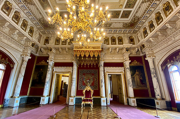 Der Thronsaal. An den Wänden befinden sich Säulen und hohe Gemälde. An der Decke hängt ein großer Kronleuchter. Decke und Wände sind mit Stuck, Bildern und Ornamenten verziert. In der Mitte des Raumes steht ein roter Samtstuhl mit Goldverzierung.