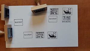 Ein weißes Blatt. Darauf liegen drei Holzstempel mit Griff. Auf dem Blatt befinden sich acht Stempelbilder in Briefmarkengröße.