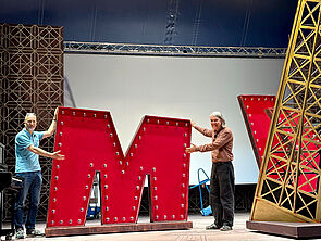 Zwei Männer stehen auf der Bühne. In ihrer Mitte ist eine Requisite. Die Requisiten ist ein großes rotes M.