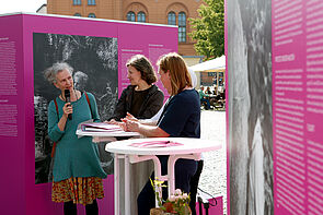Drei Frauen stehen an einem Tisch. Eine fragt, zwei antworten.