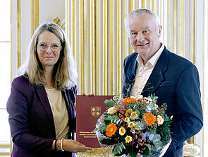 Eine Frau überreicht einem Mann eine Urkunde und einen Blumenstrauß.