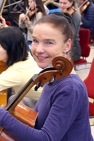 Kathrin Vogler-Georgi lächelt. An ihrer Schulter lehnt ihr Chello. Sie sitzt zwischen anderen Musikern.