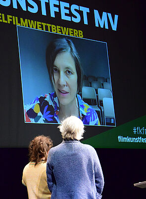 Eine Videoleinwand. Sie zeigt eine Frau. Vor der Leinwand stehen zwei Menschen. Sie schauen auf die Leinwand.