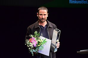 Ein Mann. Er hat einen Strauß Blumen in der Hand. Und eine Trophäe.