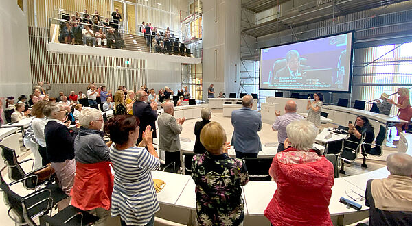 Mehrere Menschen stehen und sitzen an zu einem Rund angeordneten weißen Tischen. Sie applaudieren und zeigen fröhliche Gesichter. Im Hintergrund werden an einer Leinwand Menschen von der Live-Übertragung der Welterbeverkündung aus Indien gezeigt.