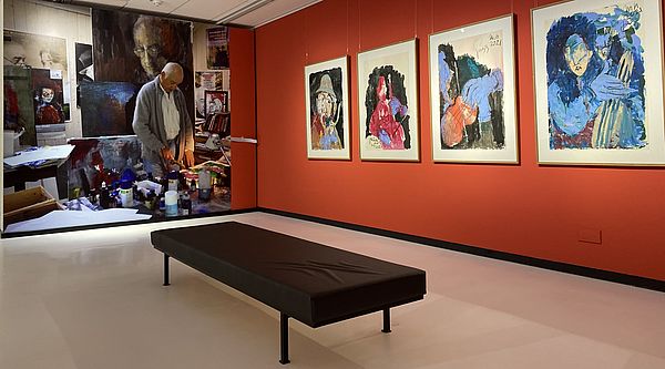 An einer roten Wand hängen vier quadratische Bilder. Daneben befindet sich ein Wandhohes und Wandbreites Foto von Armin Mueller-Stahl. Es zeigt den Künstler in seinem Atelier.