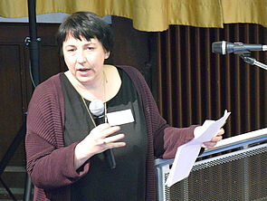 Katerina Schumacher hält ein Mikrofon in der Hand und spricht in den Saal.