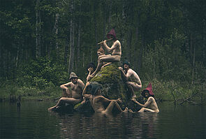 Sieben Frauen und Männer sitzen nackt im Wasser und auf einem Baumstumpf. Alle Leute tragen Hüte. 