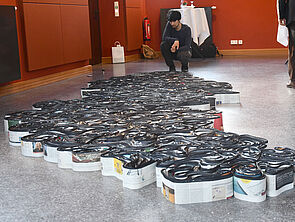 Zeitungen liegen als abstraktes Gebilde auf dem Boden. Vor ihnen hockt ein Mann.