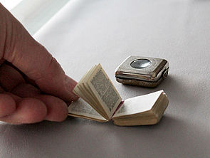 Eine Hand hält ein geöffnetes Buch. Es ist etwas kleiner als eine Streichholzschachtel. Jede Seite enthält zwei Spalten Text. Im Hintergrund liegt eine silberne Metalldose mit einer lupenartigen Einfassung. In der Dose wird das Buch aufbewahrt. 