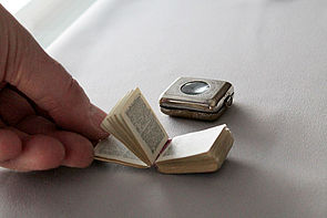 Eine Hand hält ein geöffnetes Buch. Es ist etwas kleiner als eine Streichholzschachtel. Jede Seite enthält zwei Spalten Text. Im Hintergrund liegt eine silberne Metalldose mit einer lupenartigen Einfassung. In der Dose wird das Buch aufbewahrt. 