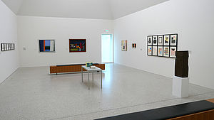 Ein weißer Raum. An den Wänden hängen Bilder. In der Ecke steht eine Skulptur. In der Mitte befinden sich eine Sitzbank und ein weißer Tisch.