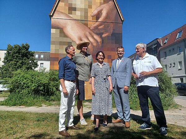 Gruppenbild mit fünf Personen. Im Hintergrund ist ein riesiges Wandbild zu sehen. Motiv: zwei Hände.