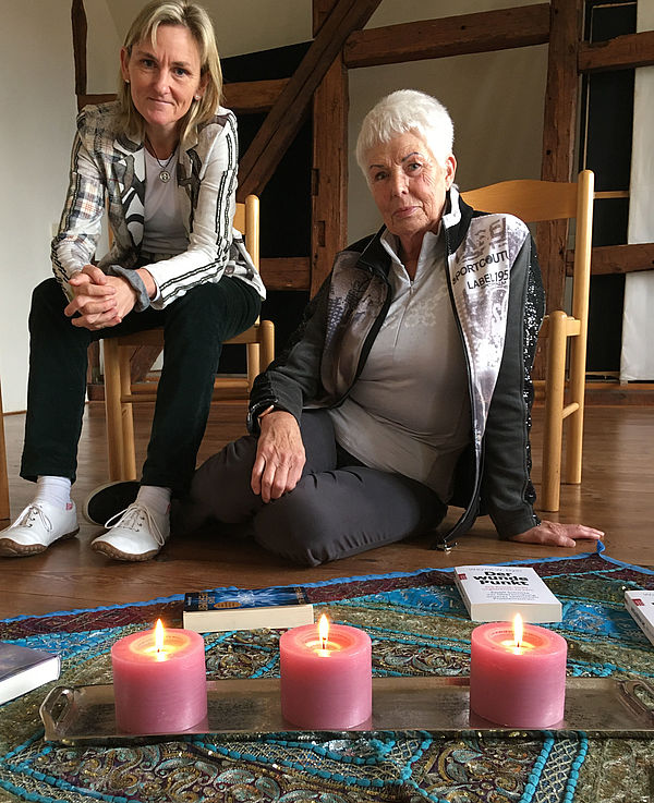 Liana Döscher und Anke Erika Thöm sitzen auf bzw. vor einem Stuhl. Auf dem Boden vor ihnen liegt ein Tuch. Darauf stehen drei Kerzen und liegen Bücher. 