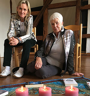 Liana Döscher und Anke Erika Thöm sitzen auf bzw. vor einem Stuhl. Auf dem Boden vor ihnen liegt ein Tuch. Darauf stehen drei Kerzen und liegen Bücher. 