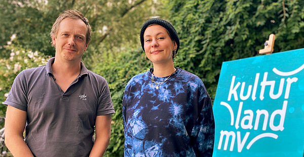 Hendrik Menzo und Betty Koschka vor einer blauen Leinwand mit dem weißen Schriftzug von Kulturland MV.