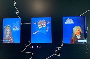 Drei blaue Bildschirme. Links und rechts darauf: zwei Monarchen. In der Mitte Herrschaftswappen.