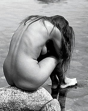 Eine Frau sitzt mit rundem Rücken nackt auf einem Stein im flachen Wasser.