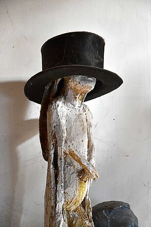 Das Foto zeigt eine weiße Skulptur. Die Skulptur trägt einen schwarzen Hut. Hans W. Scheibner hat die Skulptur gemacht. Eine Skulptur ist eine Figur aus Stein oder Ton.
