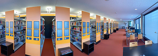 Links stehen mehrere Regale mit Büchern. Rechts Tische mit Stühlen und Computern, an denen die Bibliotheksnutzerinnen und -nutzer arbeiten können.