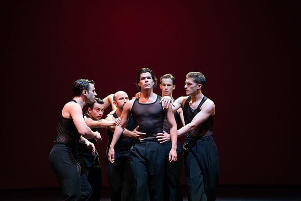 Sechs Tänzer stehen eng beieinander auf einer dunklen Bühne.