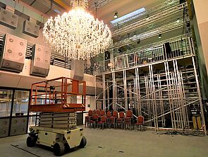 Im Raum steht ein Gabelstapler. An der Decke hängt ein Kronleuchter. An der Wand stehen rote Stühle und ein Gerüst.