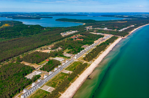 Prora von oben. Neben dem Ostseestrand wächst Grün. Daneben reiht sich ein Block an den nächsten. Dahinter befindet sich Wald. Wald, Häuserblöcke und Wasser reichen bis zum Horizont.