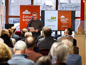 Ein Mann steht an einem Mikrofon und spricht. Vor ihm sitzen viele Leute. Hinter ihm hängen Banner zum Stadtjubiläum und vom Zukunftsschloss.