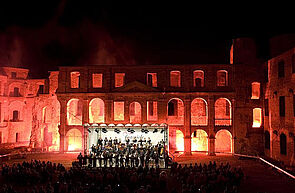 Die Ruine bei Dunkelheit. Vor der Ruine spielt ein Orchester auf einer Bühne. Viele Leute schauen zu. Die Ruine ist angestrahlt und erscheint in rötlichem Licht.