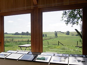 Vor einem großen Fenster stehen Tische. Darauf liegen Bilderrahmen mit Fotografien. Draußen befinden sich Hügel, Wiesen und Bäume.