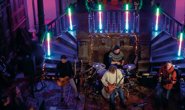 Ein dunkler Raum. Vier Musiker stehen auf einer Bühne. im Hintergrund sind viele Treppen. Der Raum ist dunkel. Scheinwerfer setzen rote und lila Lichtpunkte. Vor der Bühne stehen Zuschauer.