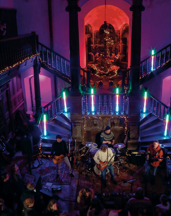 Ein dunkler Raum. Vier Musiker stehen auf einer Bühne. im Hintergrund sind viele Treppen. Der Raum ist dunkel. Scheinwerfer setzen rote und lila Lichtpunkte. Vor der Bühne stehen Zuschauer.